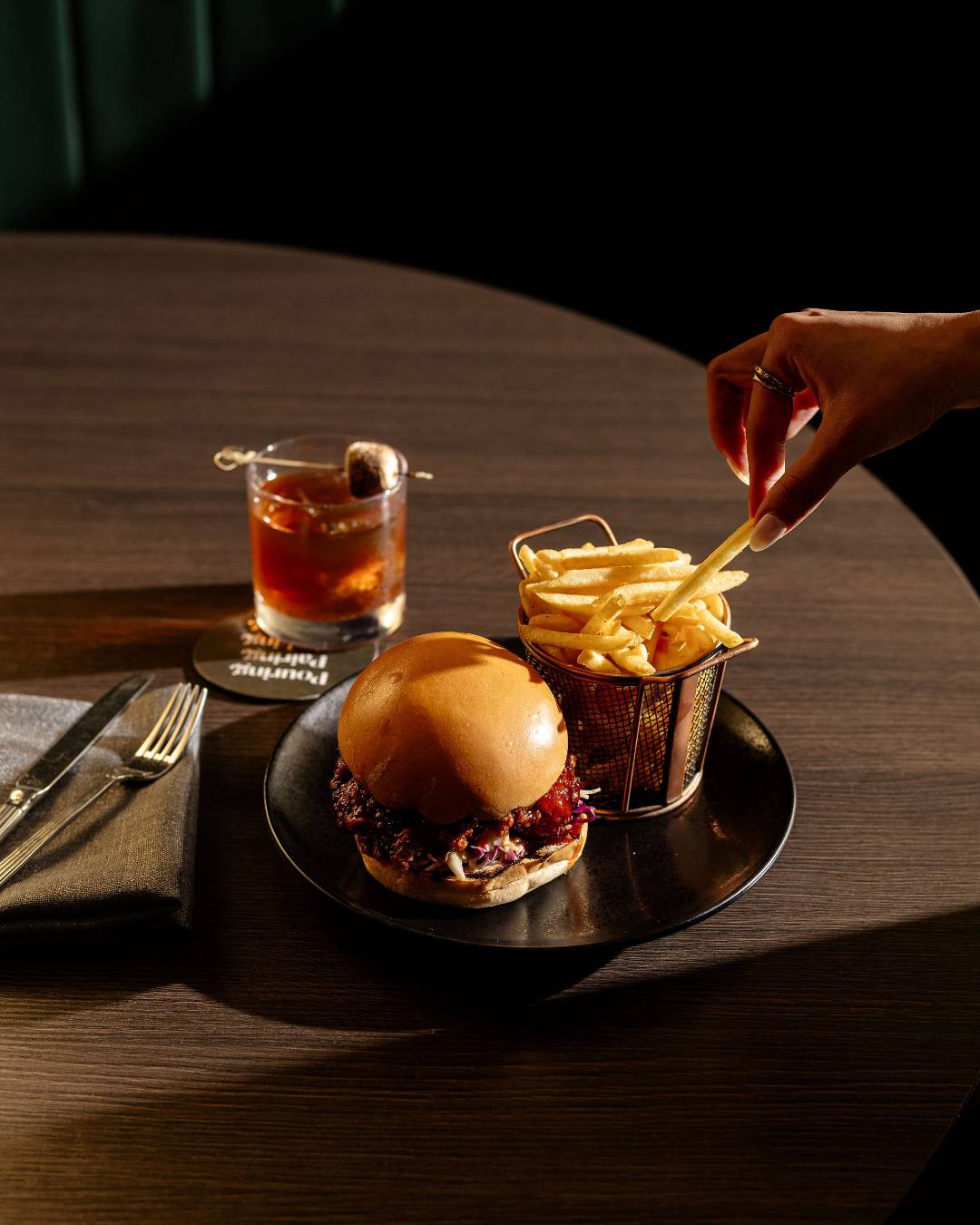 Someone picking up chips from a plate of burger and chips, cocktail on the side, from Midnight Bar in Canberra.