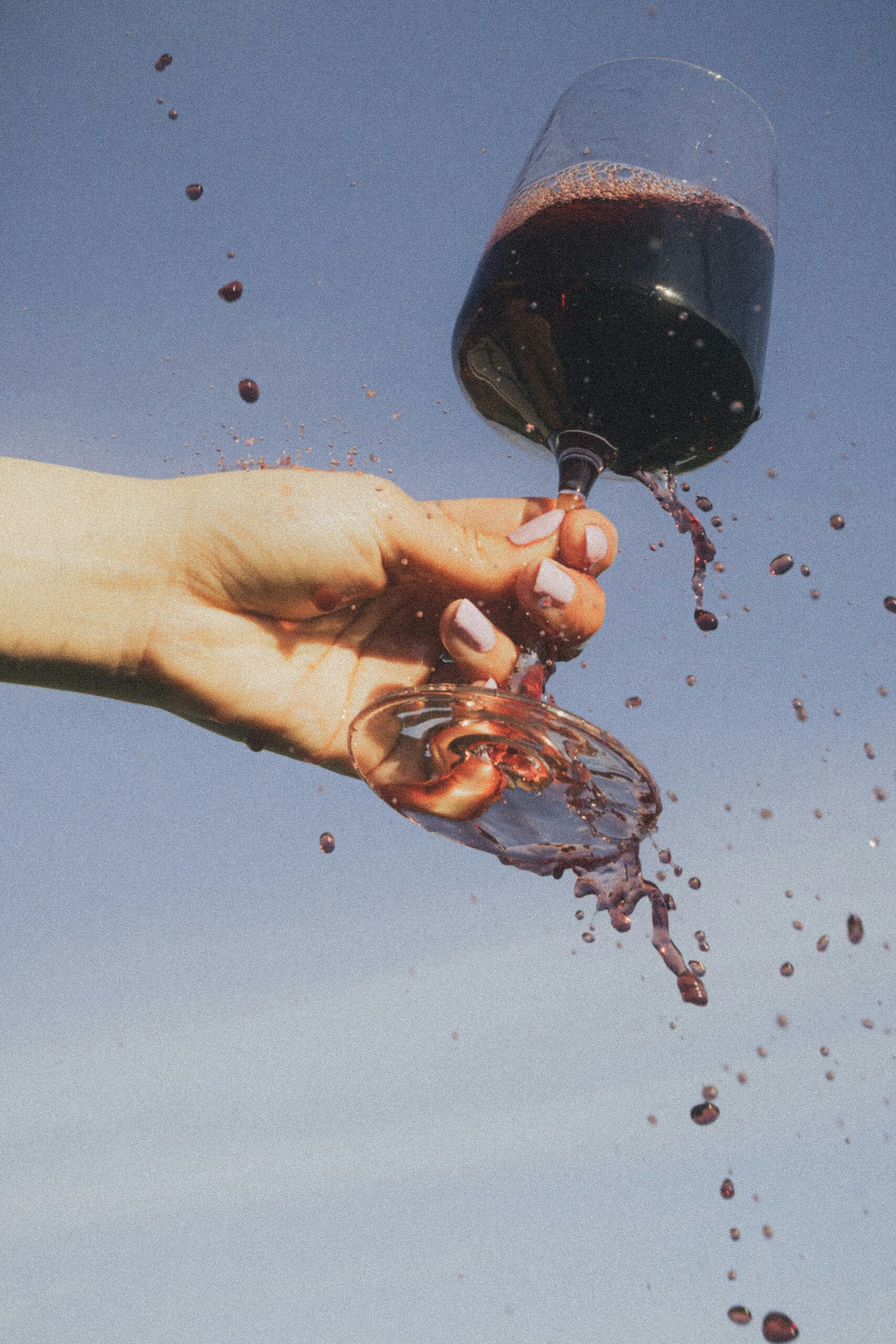 A hand holding a tilted glass of red wine as droplets splash against a blue sky.