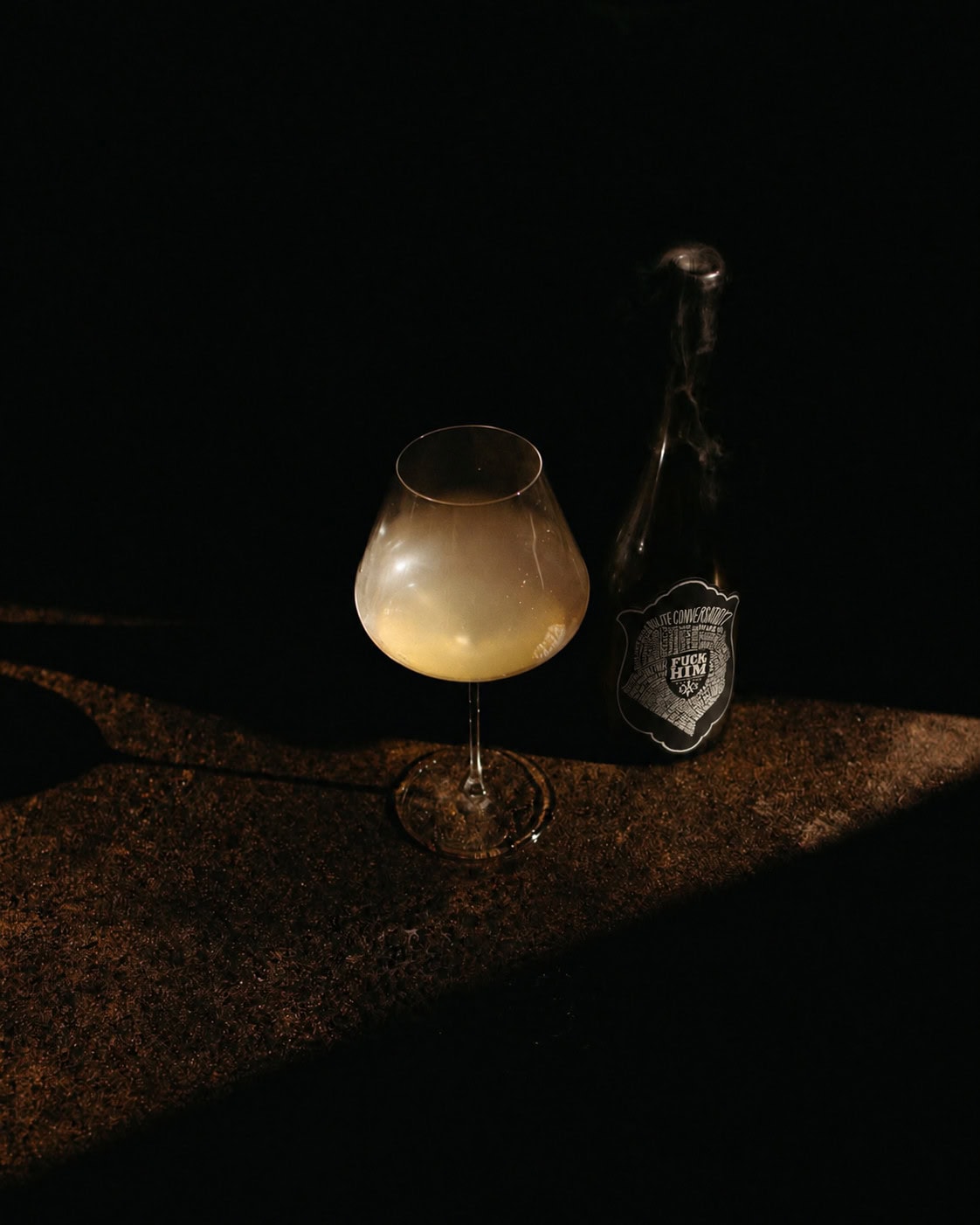 Bottle of L.A.S. Vino ‘Fuck Him’ Chardonnay beside a glass of cloudy white wine in low light.