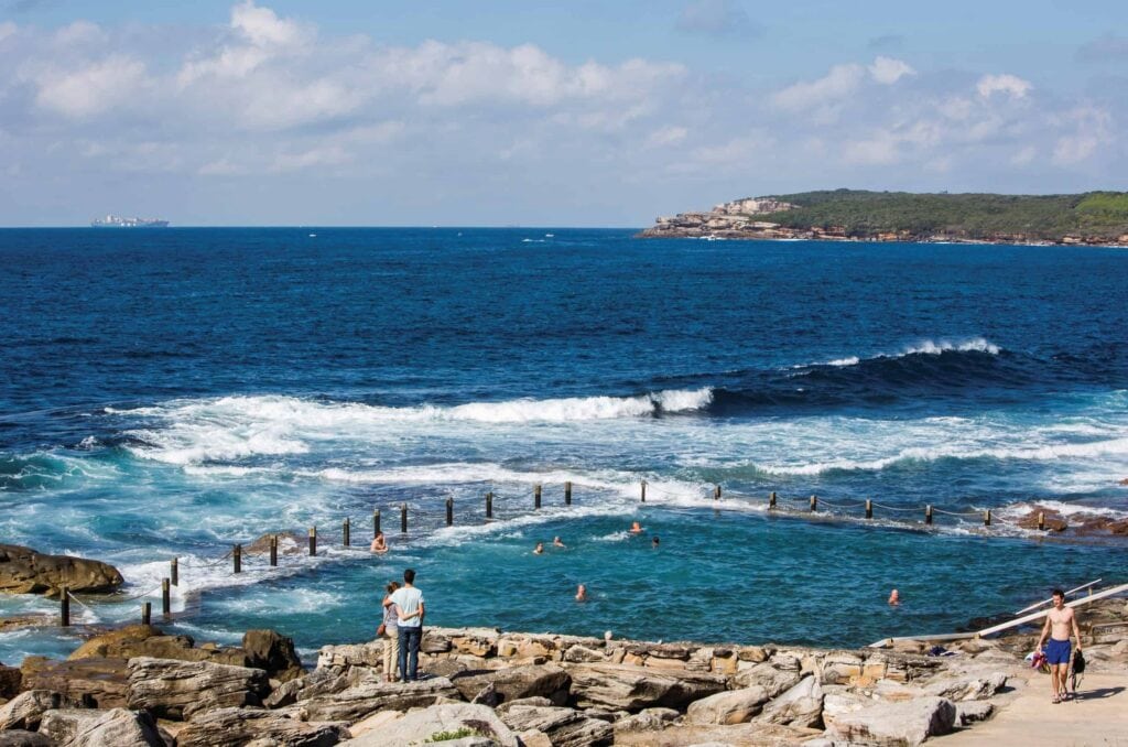 Swimmers in the ocean rock pool with waves crashing over the wall at Mahon Pool, Maroubra, Sydney