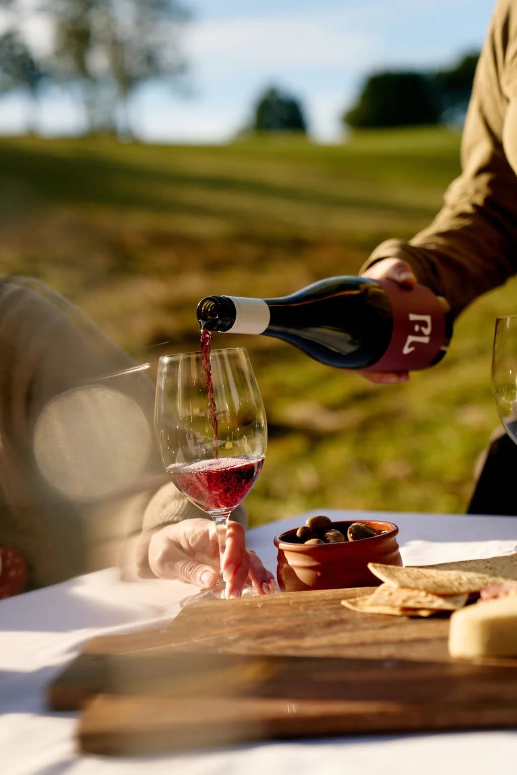 Red wine being poured into a glass at an outdoor table beside olives, crackers and cheese.