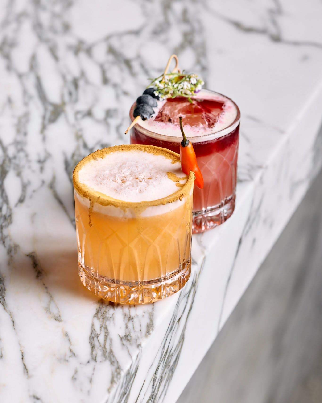 An orange and a pink cocktail in short glasses on the edge of a white marble bar.