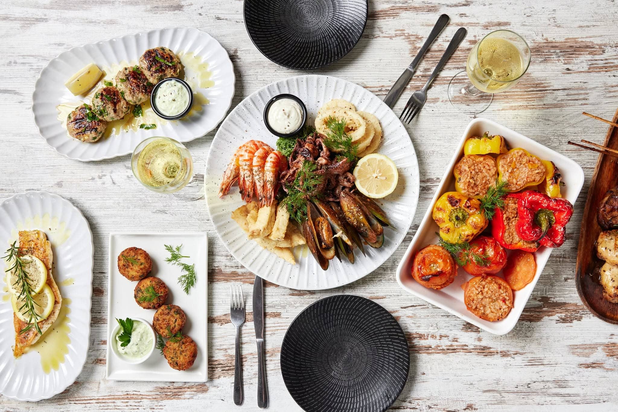A white washed table with shared plates of seafood, stuffed capsicum, meat rissoles, fish and wine.