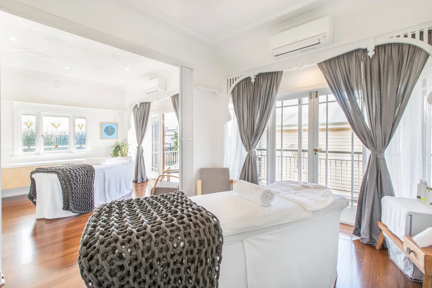 A spacious, light-filled room with two massage tables with white linens and greay decorative blankets.