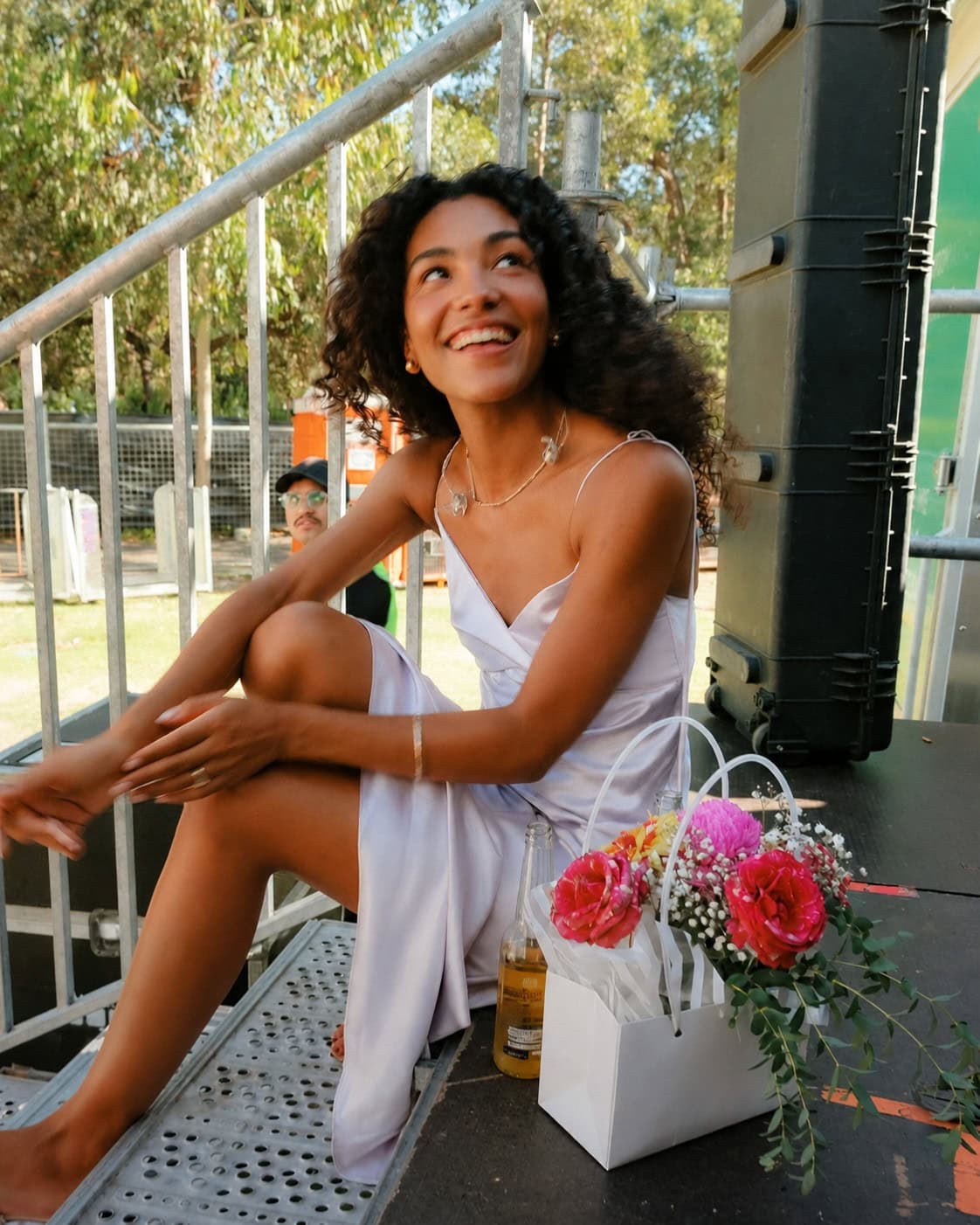 Olivia Dean sitting down in a dress on some steps with a bag filled with flowers.