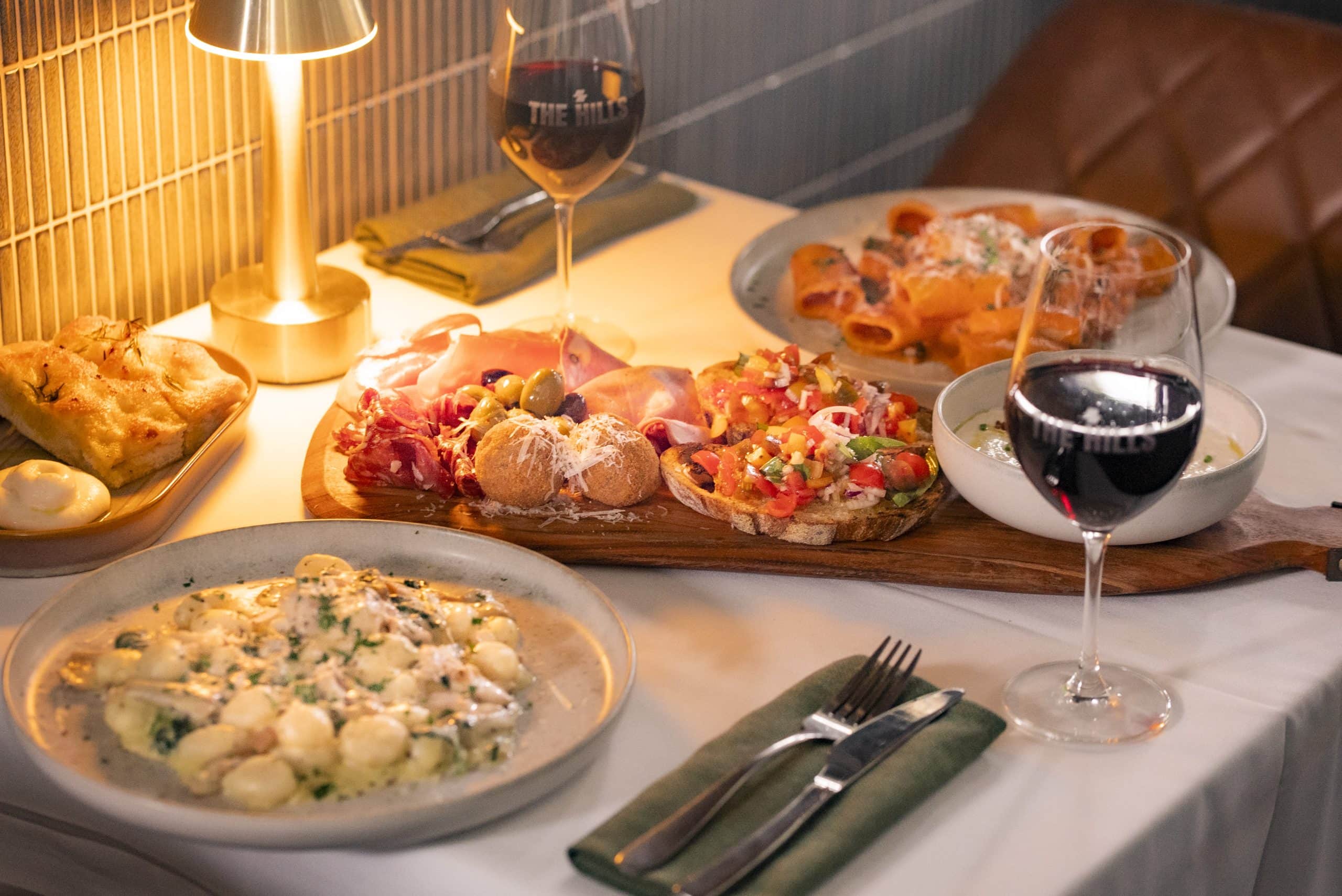 A moody lamplit table set for two, with a bowl of pasta each, a charcuterie board between them and two glasses of wine.