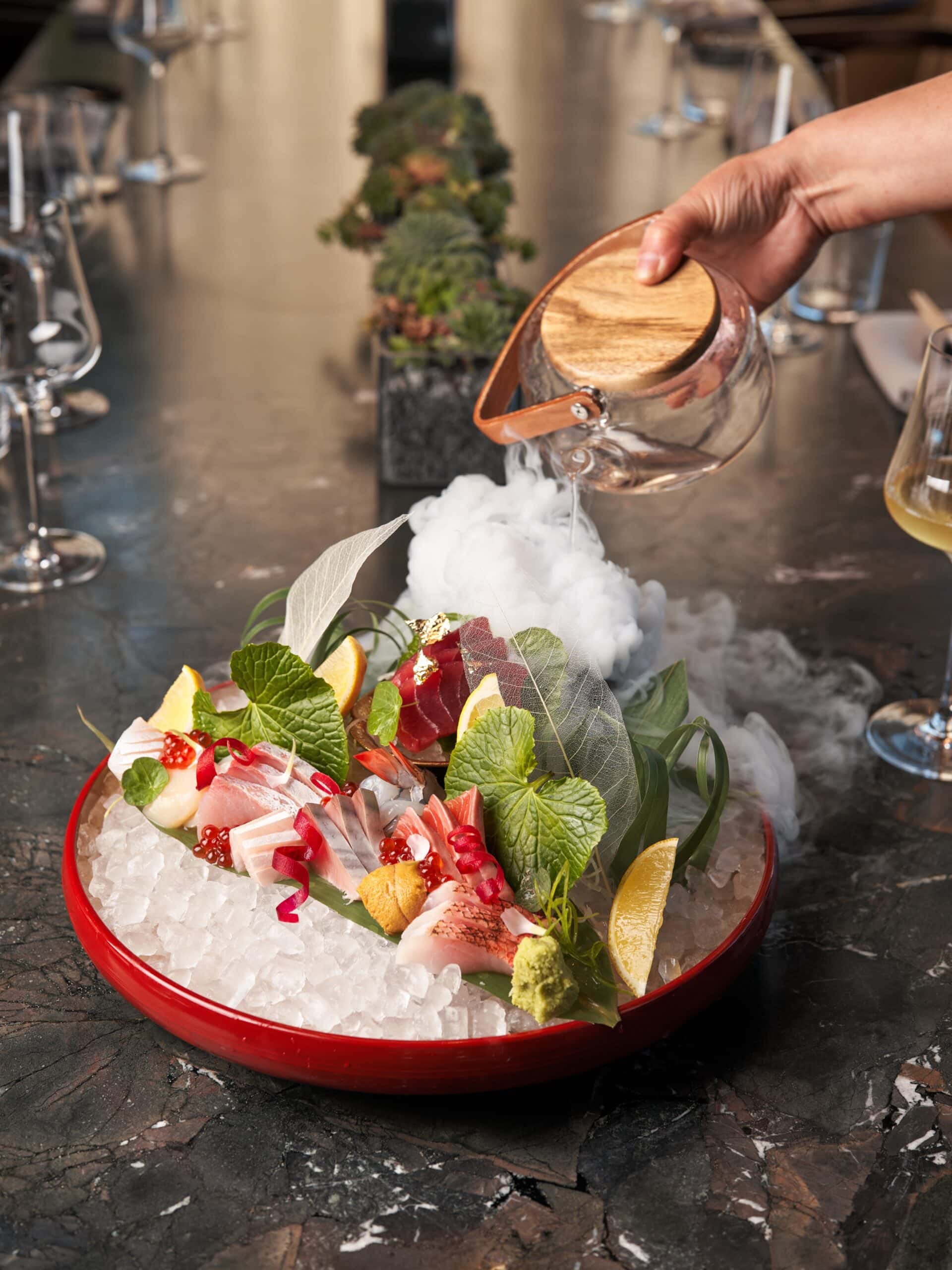 A large platter of sashimi served over ice as a hand pours dry ice over the top.