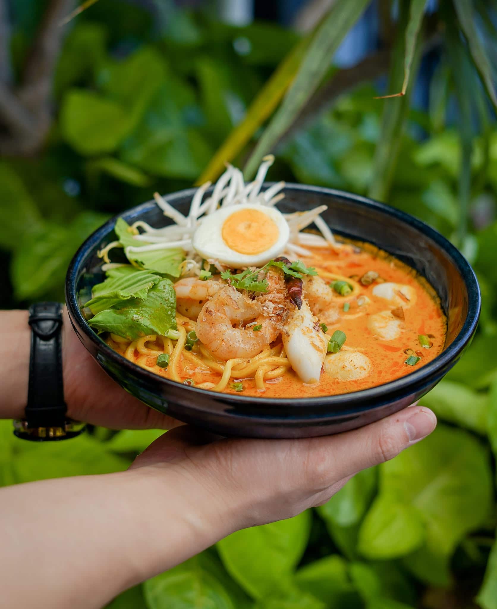 Two hands holding a black bowl of seafood laksa.