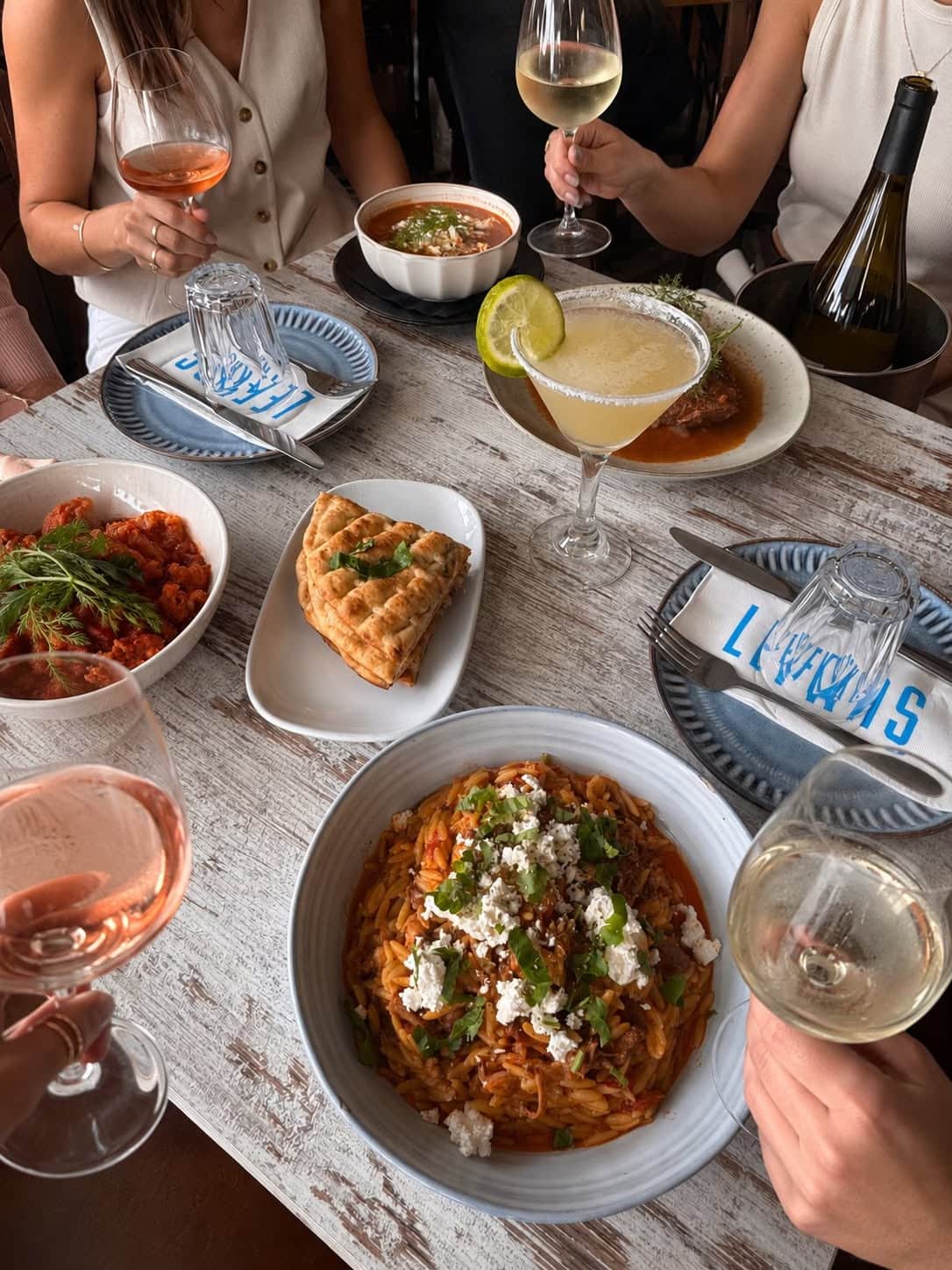 Four people at a whitewashed table with wine glasses in hand, sharing bread, orzo and a meat dish.