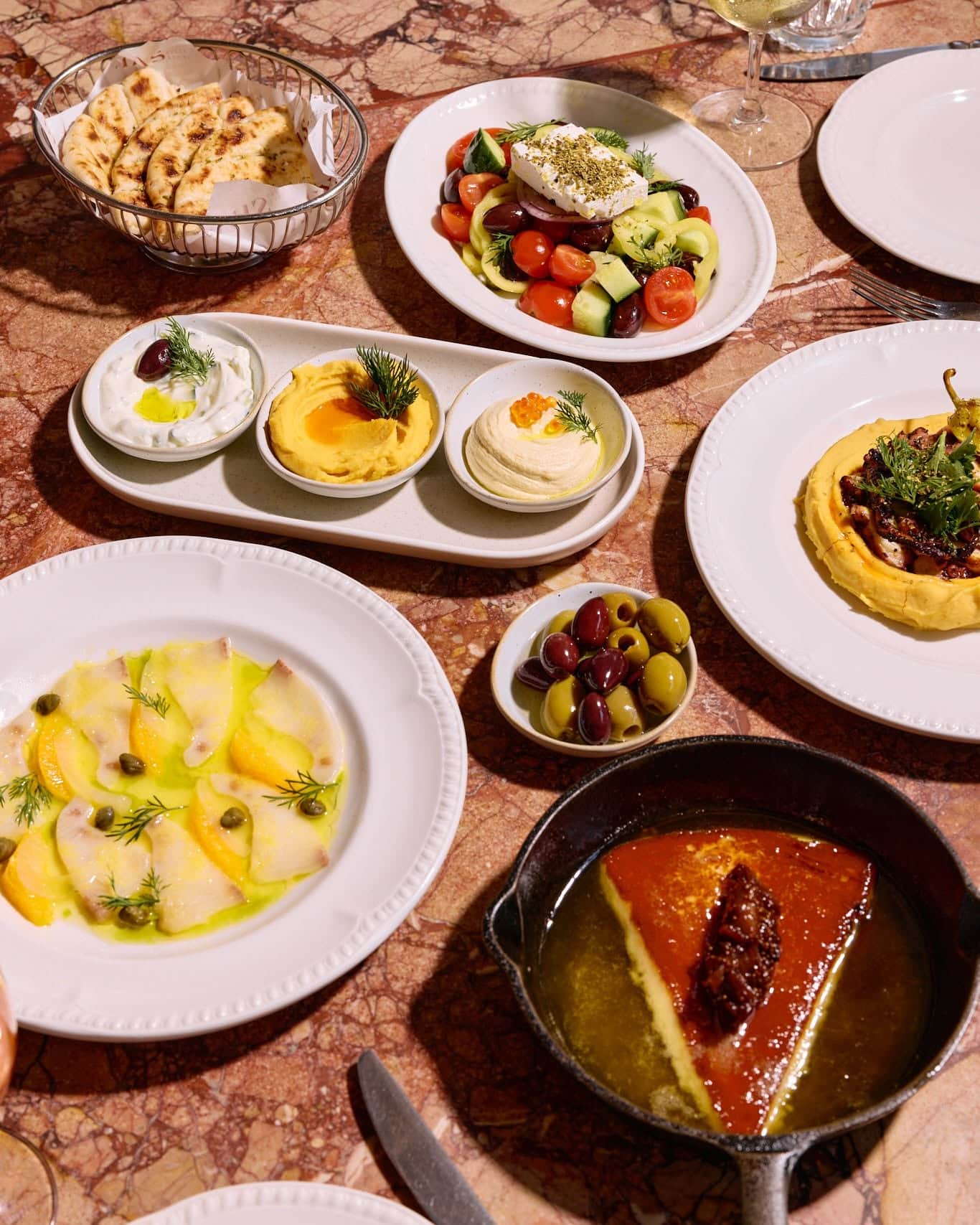 Share plates on a marble table, spanning ceviche, saganaki, olives, dips and Greek salad.