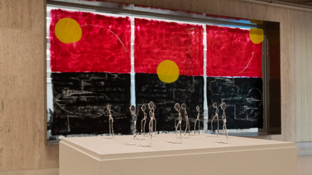 Glass figure sculptures in front of painted window panels in the colours of the Aboriginal flag, The Aboriginal Robot by Mia Khin Boe at Canberra Art Biennial 2026