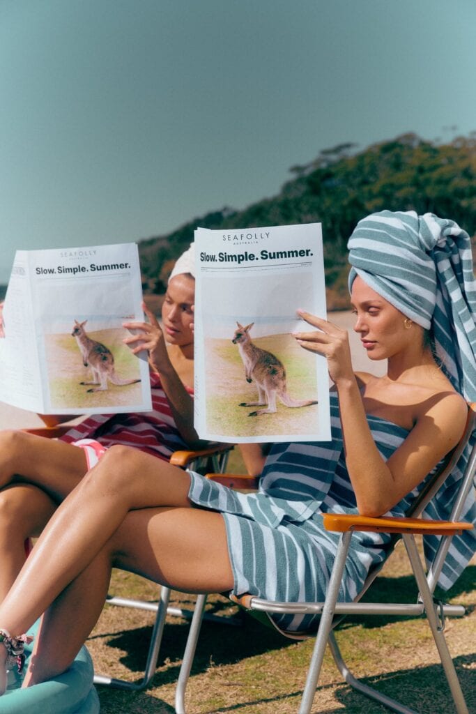 Two women wrapped in Seafolly Sunbaked and Cornflower striped terry towels reading Slow Simple Summer newspapers
