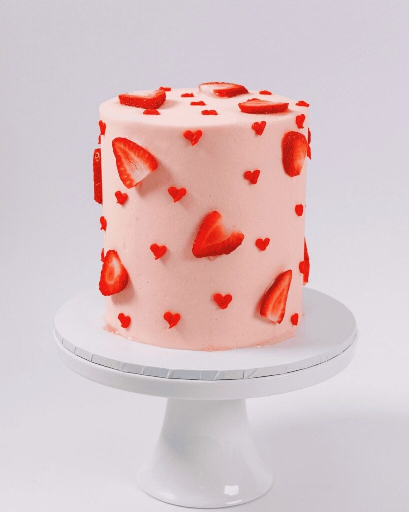 Pink buttercream layer cake decorated with fresh strawberry slices and piped red hearts on a white cake stand, by Black Velvet cake shop, Darlinghurst Sydney