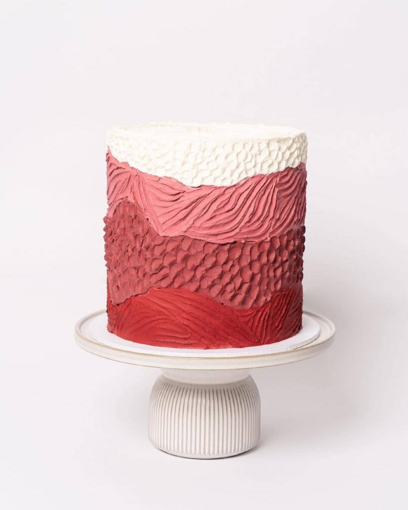 Tall layer cake with hand-textured ombre buttercream in white, pink and red on a ceramic cake stand, by online cake shop Buttercream Bakery, Sydney